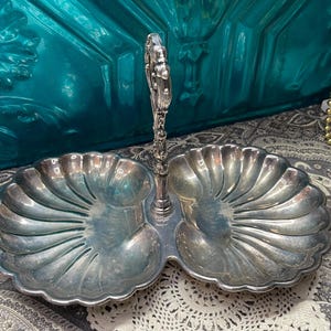 May include: A silver-toned, two-section serving dish with a handle. Each section has a scalloped, fan-like design. The handle is ornate with a floral motif. The dish is likely made of metal and is suitable for serving snacks or condiments.