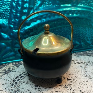 May include: A small, black, antique sugar bowl with a brass lid and handle. The lid has a small opening for a spoon. The bowl sits on three small feet. The handle is arched and attached to the sides of the bowl.