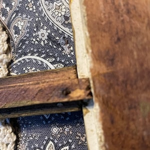 K&ouml;nnte beinhalten: Nahaufnahme eines Holzrahmens mit dunkelbrauner Oberfl&auml;che. Der Rahmen ist an einem Stoffhintergrund mit Paisley-Muster befestigt.