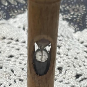 May include: A vertical wooden hourglass featuring a cylindrical body with a smooth, light brown finish. The center of the wood frame has an oval cutout revealing a clear glass bulb containing dark sand, positioned between a flat top and base.