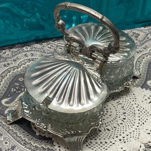 May include: A silver-colored, two-part serving dish with a handle. The dish has a shell-like design on the lids and a decorative base. The dish is open, revealing the interior.