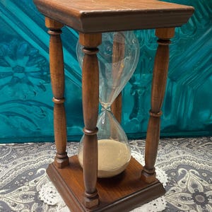 May include: A wooden hourglass with a clear glass bulb filled with sand. The hourglass is supported by a square wooden base and top, connected by four turned wooden columns. The hourglass is approximately 7 inches tall.