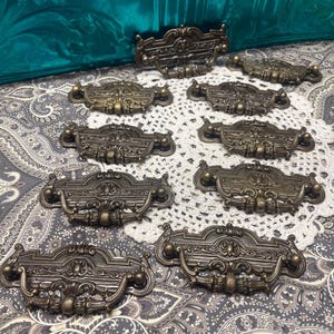 May include: A collection of antique bronze drawer pulls with ornate detailing. The pulls have a curved design with decorative elements, perfect for vintage furniture restoration. The hardware is arranged on a patterned surface.
