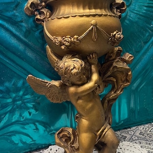 May include: A gold-colored wall sconce featuring a cherub figure supporting a decorative urn. The urn has ornate detailing, including floral accents and a scalloped rim. The cherub has detailed wings and is posed in a lifting position.