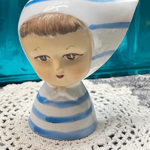 May include: A vintage ceramic figurine of a child wearing a white bonnet with blue stripes and a matching striped top. The figurine has a painted face with brown hair and delicate features. The base is a rounded shape.
