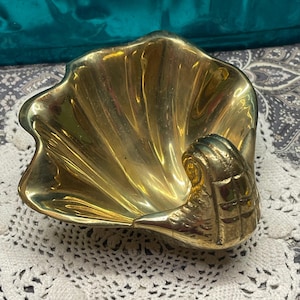 May include: A golden brass seashell-shaped dish. The dish has a textured, wavy design and a detailed seashell accent on the side. The interior of the dish reflects light, creating a shiny appearance. This decorative piece is suitable for holding small items.