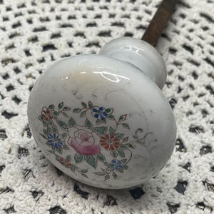 May include: A white ceramic door knob with a rounded shape featuring a central floral pattern of pink, red, and blue flowers with green leaves. A dark, rusted metal spindle extends from the back of the knob, which has a smooth, glossy finish.