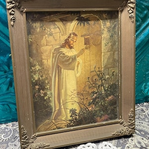 May include: A framed religious print featuring a figure in a white robe knocking on a wooden door. The print has a gold-colored frame with ornate corner details. The artwork depicts a scene with plants and a stone wall.