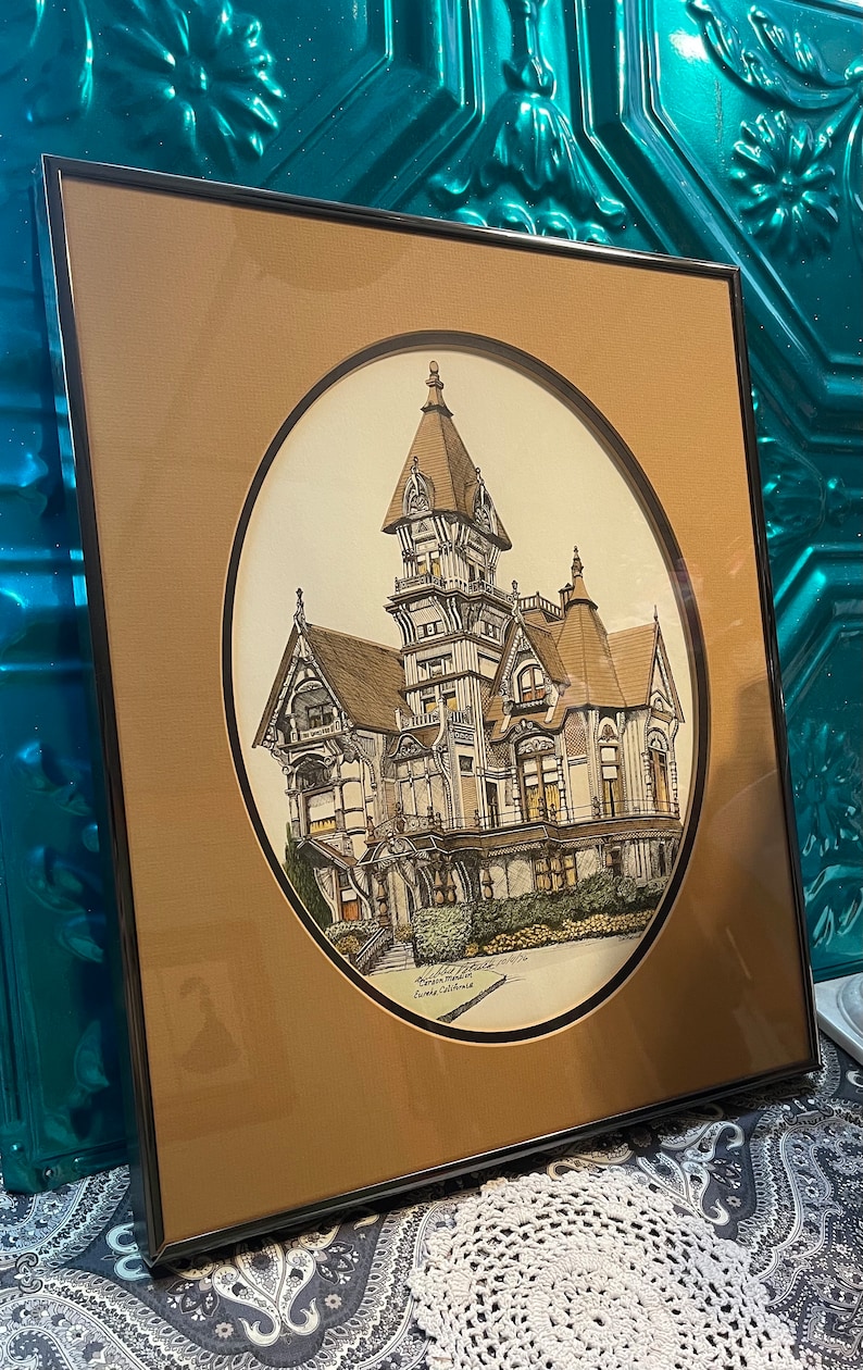 May include: A framed print of a detailed drawing of a large, ornate Victorian house with a tower. The house is painted in shades of brown, gray, and white. The print is set against a brown background and has a white oval border. The text "Leland Stanford Mansion, Garden Manor, Loomis, California" is printed at the bottom of the print.