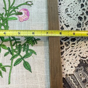 May include: A framed embroidery of a pink flower with green leaves on a linen background. The frame is made of wood and has a brown finish. A measuring tape is placed along the bottom edge of the frame.