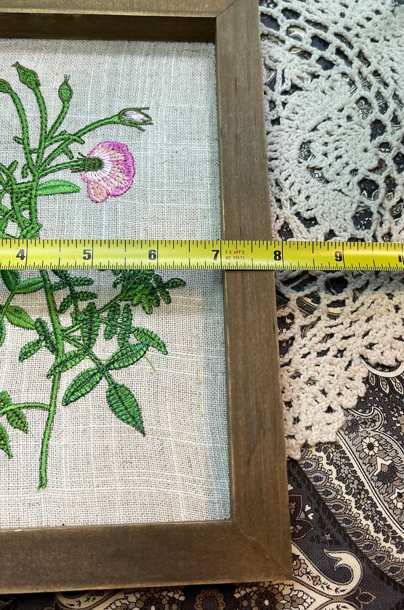 May include: A framed embroidery of a pink flower with green leaves on a linen background. The frame is made of wood and has a brown finish. A measuring tape is placed along the bottom edge of the frame.