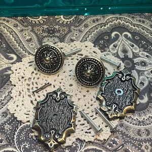 May include: Four antique-style cabinet knobs with a distressed black finish and ornate gold accents. Two knobs have a circular design with a raised floral pattern, and two knobs have a rectangular design with a textured surface.