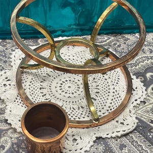 May include: A copper and brass metal trivet with a circular design. The trivet has a small copper cup next to it. The trivet is sitting on a white doily and a patterned fabric.