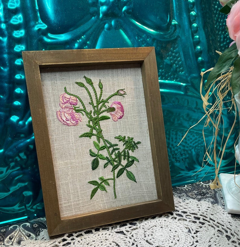 May include: A framed embroidery of pink flowers with green stems and leaves on a beige linen background.