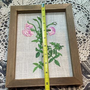 May include: A framed embroidered floral artwork featuring pink flowers and green leaves on a beige linen background. The frame is made of brown wood and measures approximately 10 inches in height.