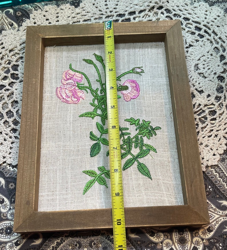 May include: A framed embroidered floral artwork featuring pink flowers and green leaves on a beige linen background. The frame is made of brown wood and measures approximately 10 inches in height.
