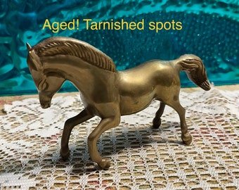 May include: Aged brass horse figurine with tarnished spots. The horse is standing on a white lace surface.