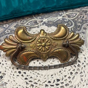 May include: Ornate antique brass drawer pull with a floral center design and curved handle. The pull has a detailed, aged appearance with visible wear and tear, adding to its vintage charm. The pull is approximately 6 inches wide.