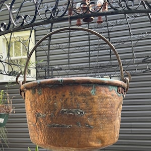 May include: A large, antique copper pot with a hammered finish hangs from a wrought iron structure. The pot has a handle and is slightly tarnished with age.