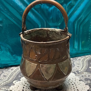 May include: A vintage copper bucket with a curved handle. The bucket features decorative silver-colored metal accents and a detailed embossed design. The bucket is resting on a white crocheted doily.