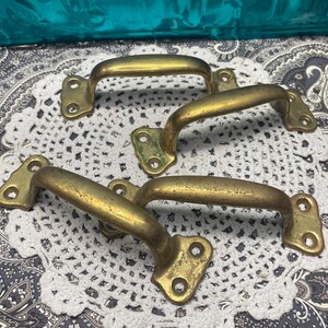 May include: A set of four brass drawer pulls, each featuring a curved handle design with two mounting plates, each containing two screw holes. The brass has a weathered, aged patina with visible surface wear and a dull, muted gold finish.