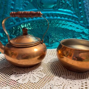 May include: A copper teapot with a wooden handle and a matching copper bowl. The teapot and bowl are sitting on a white crocheted surface.