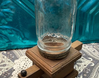 May include: A clear glass jar with a metal lid sits atop a wooden base. The base is a dark brown color and has a small wooden knob on one side. The jar is cylindrical and appears to be empty.