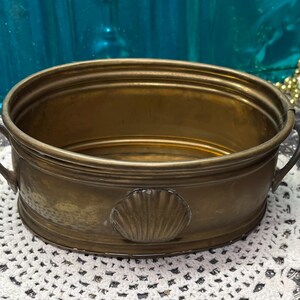 May include: An oval brass basin featuring two side handles and a decorative embossed seashell motif on the front exterior. The metal vessel has a polished finish with visible natural patina and a smooth, rounded rim, designed as a functional container.