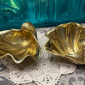 May include: Two gold-colored seashell-shaped decorative bowls. The bowls have a wavy edge and a detailed, ornate design on the side. They are placed on a white lace doily and a patterned fabric background.
