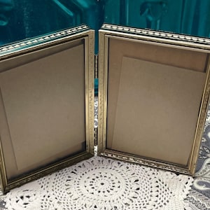 May include: A hinged, double-sided picture frame with a gold-colored frame and a decorative border. The frame is open, revealing the interior space for photos or artwork. The frame is resting on a white, circular doily.