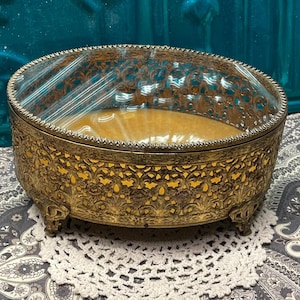 May include: An antique oval jewelry box with a clear glass lid and ornate filigree metalwork in a gold tone. The interior has a tan fabric lining. The box sits on small, decorative feet and is placed on a white lace doily.