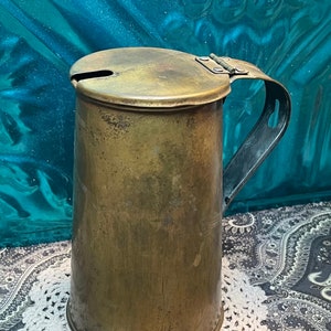 May include: A vintage brass pitcher with a hinged lid and a handle. The pitcher has a worn, aged patina and is sitting on a white crocheted doily.