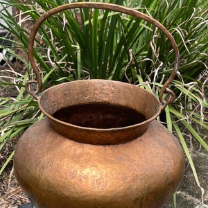 May include: A large, hammered copper pot with a curved handle. The pot has a rustic, aged appearance and is perfect for a farmhouse or rustic decor style.
