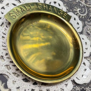 May include: A brass coin dish with the words "SMALL CHANGE" embossed on the curved top. The dish is round with a shallow bowl and sits on a white lace doily.
