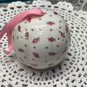 May include: A spherical white ceramic pomander featuring a repeating pattern of small pink roses and green leaves. The surface has subtle vertical ridges and small circular perforations. A light pink satin ribbon is attached to the top for hanging.