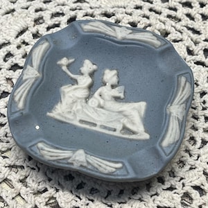 May include: A small, blue ashtray with a white bas-relief design of two figures on a sled. The ashtray has a scalloped edge with white decorative accents. A vintage piece.