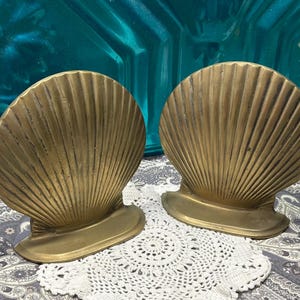 May include: Two gold-colored seashell bookends. Each bookend has a fan-shaped design with vertical lines. The bookends are on a patterned surface with a white lace doily. The background is teal.