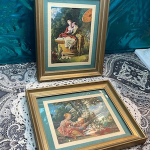 May include: Two framed vintage prints with gold frames. The top print depicts a couple in a garden setting with a parasol. The bottom print shows a couple sitting under a tree.
