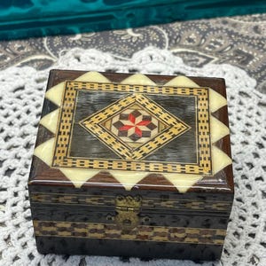 May include: A small wooden box with a geometric inlay design. The box is made of dark wood with a light wood inlay. The inlay is in the shape of a diamond and features a red star in the center. The box has a brass latch.
