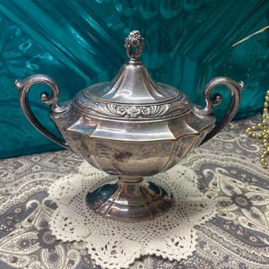 May include: A silver-colored, lidded serving dish with ornate handles and a decorative finial. The dish has a pedestal base and is resting on a white, lacy doily. The background is a teal-colored surface.