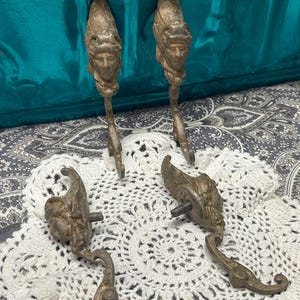 May include: Antique bronze-toned cabinet pulls with a classical face design. The pulls feature a detailed face at the top and a curved hook shape. The set includes four pieces, perfect for adding a vintage touch to furniture.