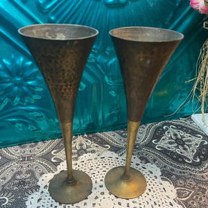 May include: Two hammered brass champagne flutes with gold-colored stems. The flutes are on a white crocheted doily.