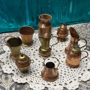 May include: A collection of miniature copper and brass kitchenware items, including a bucket, a pitcher, a teapot, and a small vase. The items are arranged on a white, crocheted doily. The pieces have a vintage aesthetic.