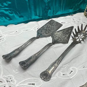 May include: Three antique silver serving utensils: a pie server, a cake server, and a spaghetti server. Each piece has ornate detailing on the handles and serving surfaces. The utensils are arranged on a white, embroidered cloth.
