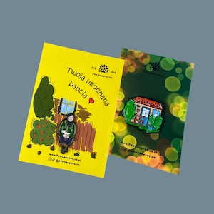 May include: Two enamel pins on cards. One pin features a house with the word "Dziadka" and the other shows a woman with a cat. The cards are yellow and green with text and illustrations. The pins are colorful.