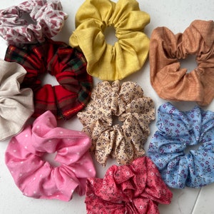 May include: A collection of nine fabric scrunchies in various colours and patterns, including pink with white polka dots, red plaid, beige with floral print, and blue with white floral print.