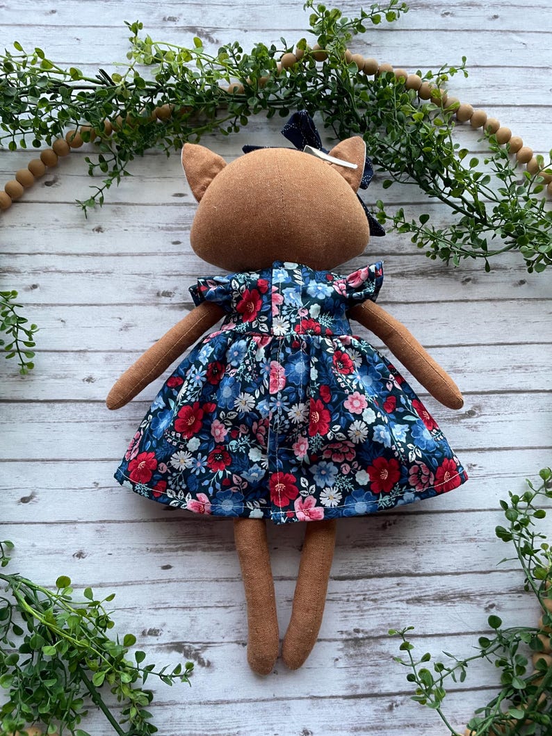 Op de afbeelding: Een stoffen pop met een bruine stoffen huid, gekleed in een donkerblauwe jurk met een levendig bloemenpatroon van rode, roze en lichtblauwe bloemen. De pop heeft een donkerblauwe stoffen strik die met een zilveren clip op het hoofd is bevestigd.