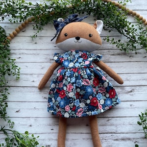 May include: A plush fox doll with brown fur and a light gray muzzle, wearing a dark blue dress featuring a vibrant floral pattern of red, pink, and light blue flowers. The fox has a dark blue bow on its head and simple embroidered facial features.