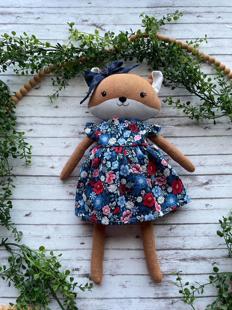 Op de afbeelding: Een pluche vossenpop met bruine vacht en een lichtgrijze snuit, gekleed in een donkerblauwe jurk met een levendig bloemenpatroon van rode, roze en lichtblauwe bloemen. De vos heeft een donkerblauwe strik op het hoofd en geborduurde details.