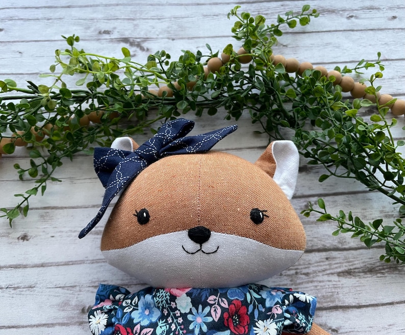 Op de afbeelding: Een knuffelvos van bruine en witte stof, met geborduurde zwarte ogen en een neus. De vos draagt een donkerblauwe stoffen strik met een wit geometrisch patroon op het hoofd en een donkerblauwe jurk met een kleurrijke bloemenprint.
